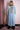 Woman wearing long light blue denim trench coat with embroidered lion design on back, standing indoors