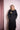 Woman wearing a black belted trench coat with decorative gold patch, standing against pink backdrop