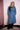 Woman wearing a long blue denim trench coat with black feather brooch, standing indoors