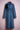 Long denim trench coat with belt and black feather brooch on pink mannequin against light pink background